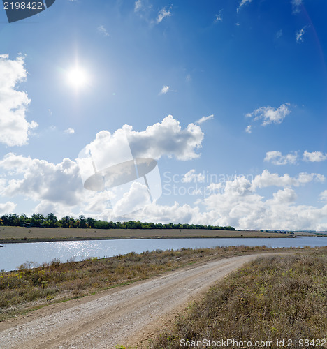 Image of sun on cloudy sky over rural road and river near it