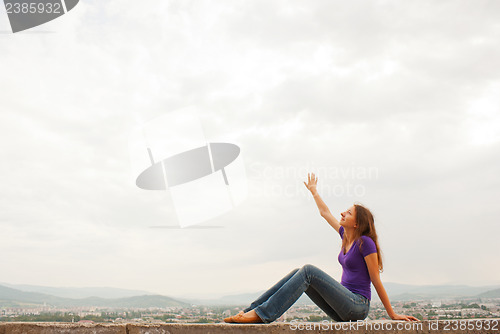 Image of Young woman sitting with raised hand against blue sky