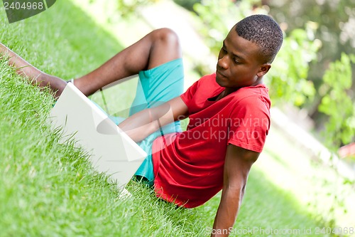 Image of young smiling african student sitting in grass with notebook