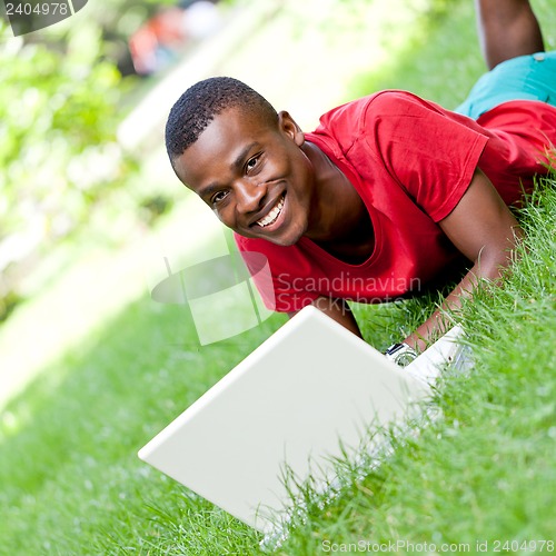 Image of young smiling african student sitting in grass with notebook