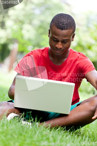 Image of young smiling african student sitting in grass with notebook