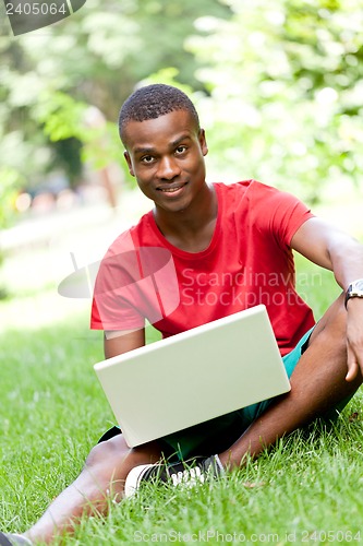 Image of young smiling african student sitting in grass with notebook