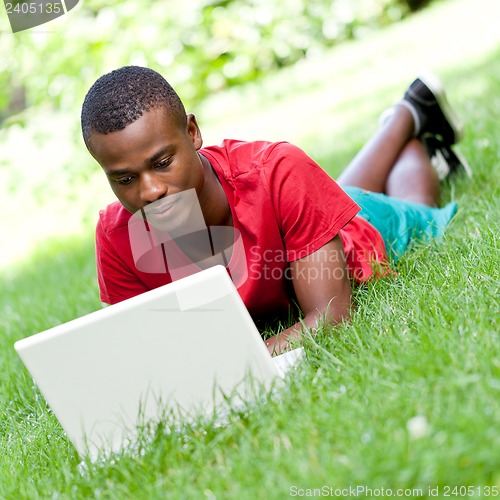 Image of young smiling african student sitting in grass with notebook