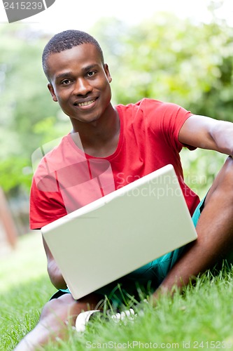 Image of young smiling african student sitting in grass with notebook