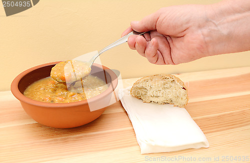 Image of Hand holds spoonful of soup with bread roll and napkin on table