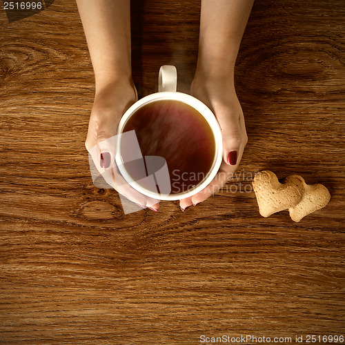 Image of woman holding hot cup of tea with cookies on wooden table