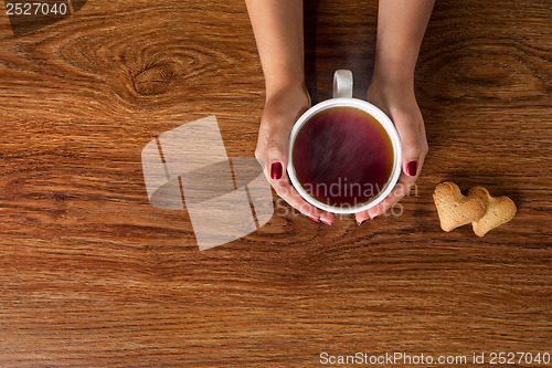 Image of woman holding hot cup of tea with cookies on wooden table