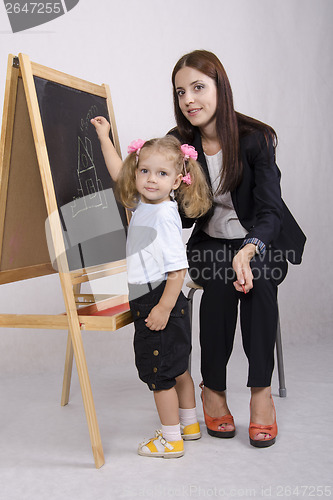 Image of Little girl draws on Board house, educator helps her