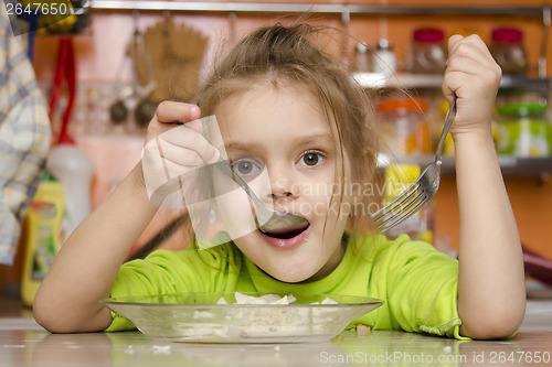 Image of girl eats with a fork and spoon sitting at table in kitchen