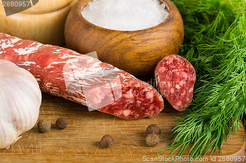 Image of sliced salame on cutting board, with dill, pepper, salt