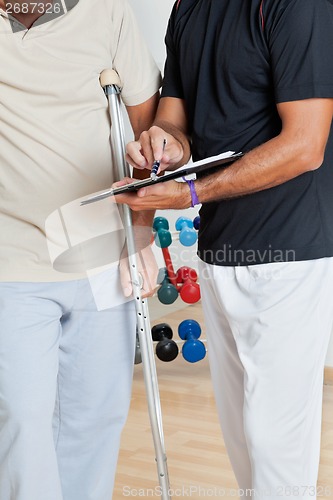 Image of Trainer Holding Clipboard While Standing With Senior Man On Crut