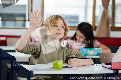 Image of Schoolboy Looking Away While Raising Hand At Desk