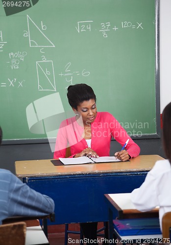 Image of Teacher Writing In Binder At Desk With Students In Foreground