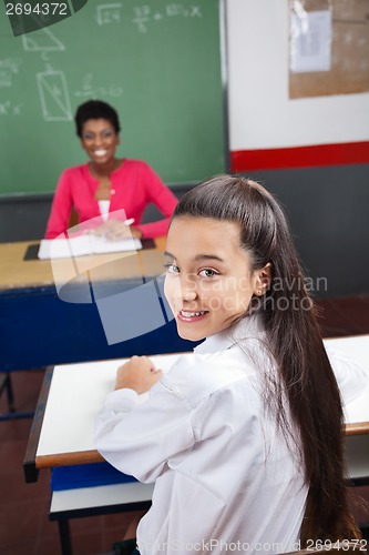 Image of Schoolgirl Sitting At Desk With Teacher Smiling In Background