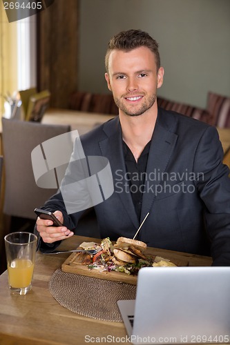 Image of Businessman With Mobilephone And Laptop Having Meal In Restauran