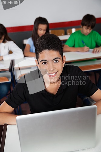 Image of Happy Teenage Schoolboy Sitting With Laptop In Classroom