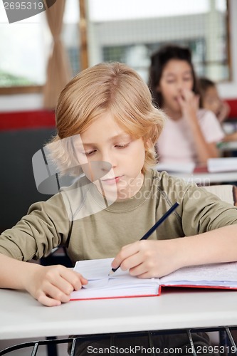 Image of Cute Schoolboy Writing In Book At Desk