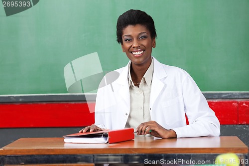 Image of Happy Teacher Sitting With Binder At Desk In Classroom