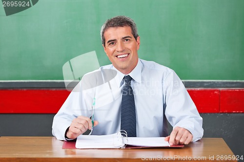 Image of Happy Male Teacher With Pen And Binder Sitting At Desk