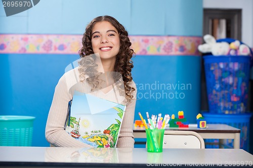 Image of Teacher With Book Sitting At Desk In Kindergarten