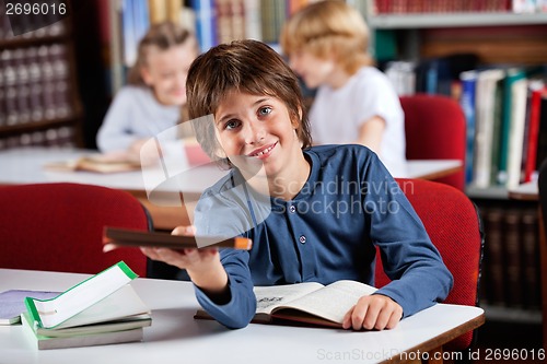Image of Cute Schoolboy Giving Book While Sitting At Table In Library