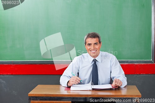 Image of Confident Male Teacher With Pen And Binder Sitting At Desk