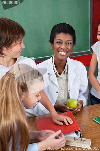 Image of Teacher Holding Apple With Students Standing At Desk