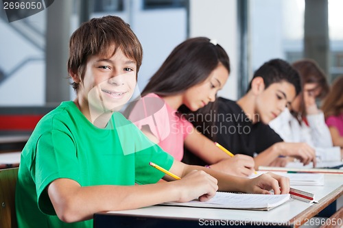 Image of Boy Sitting At Desk With Friends Writing In Classroom