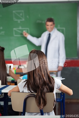 Image of Girl Sitting At Desk With Teacher Teaching In Background