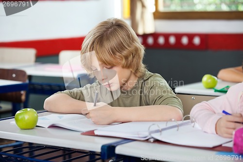 Image of Schoolboy Writing On Book In Classroom