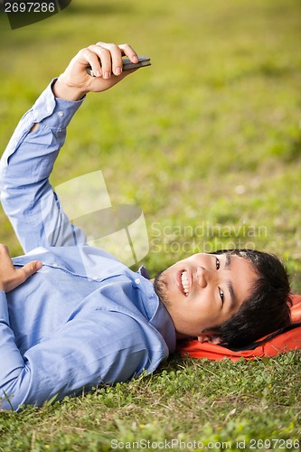 Image of College Student Holding Mobilephone While Lying On Grass At Camp