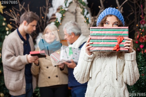 Image of Thoughtful Woman Holding Christmas Present With Family In Backgr