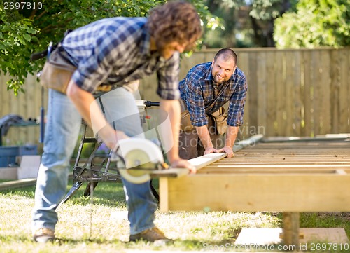 Image of Carpenter Looking At Coworker While Assisting Him In Cutting Woo