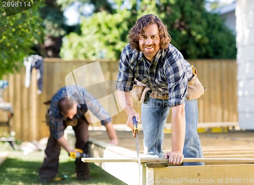 Image of Carpenter Cutting Wood With Saw While Coworker Drilling At Site