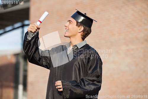 Image of Man In Graduation Gown Looking At Certificate On Campus