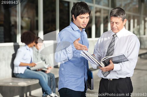 Image of Teacher Holding Books While Discussing With Student On Campus