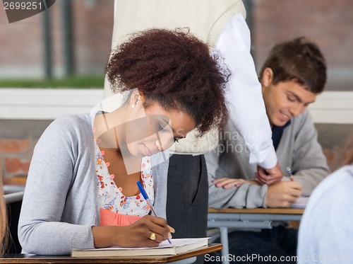 Image of Student Writing Exam While Teacher Explaining Classmate In Backg