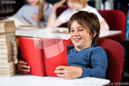 Image of Schoolboy Smiling While Reading Book At Table In Library