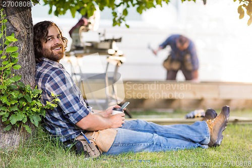 Image of Carpenter Holding Mobilephone And Cup While Leaning On Tree Trun