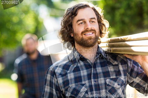 Image of Carpenter Smiling While Carrying Planks With The Help Of Coworke