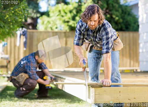 Image of Worker Cutting Wood With Saw While Coworker Drilling At Site