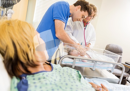 Image of Mature Doctor Examining Newborn Babygirl While Couple Looking At