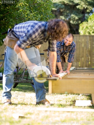 Image of Carpenter Assisting Coworker In Cutting Wood With Handheld Saw