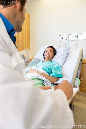 Image of Patient Looking At Doctor Writing On Clipboard On Hospital Bed