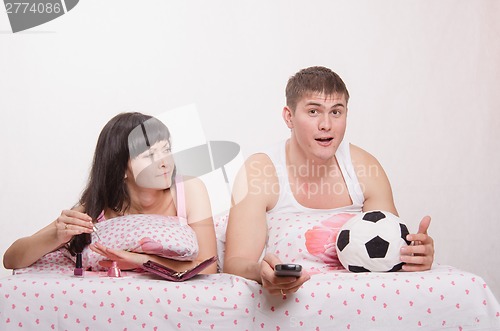 Image of Girl paints her nails in bed, man emotionally watching soccer