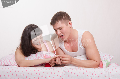 Image of Guy puts a ring on finger of girl in bed