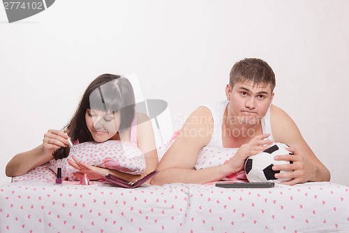 Image of Girl paints her nails in bed, man emotionally watching soccer