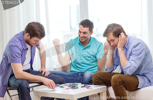 Image of happy three male friends playing poker at home