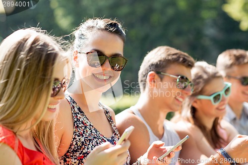 Image of smiling friends with smartphones sitting in park