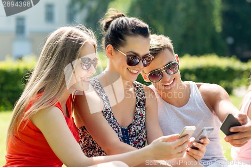 Image of smiling friends with smartphones sitting in park